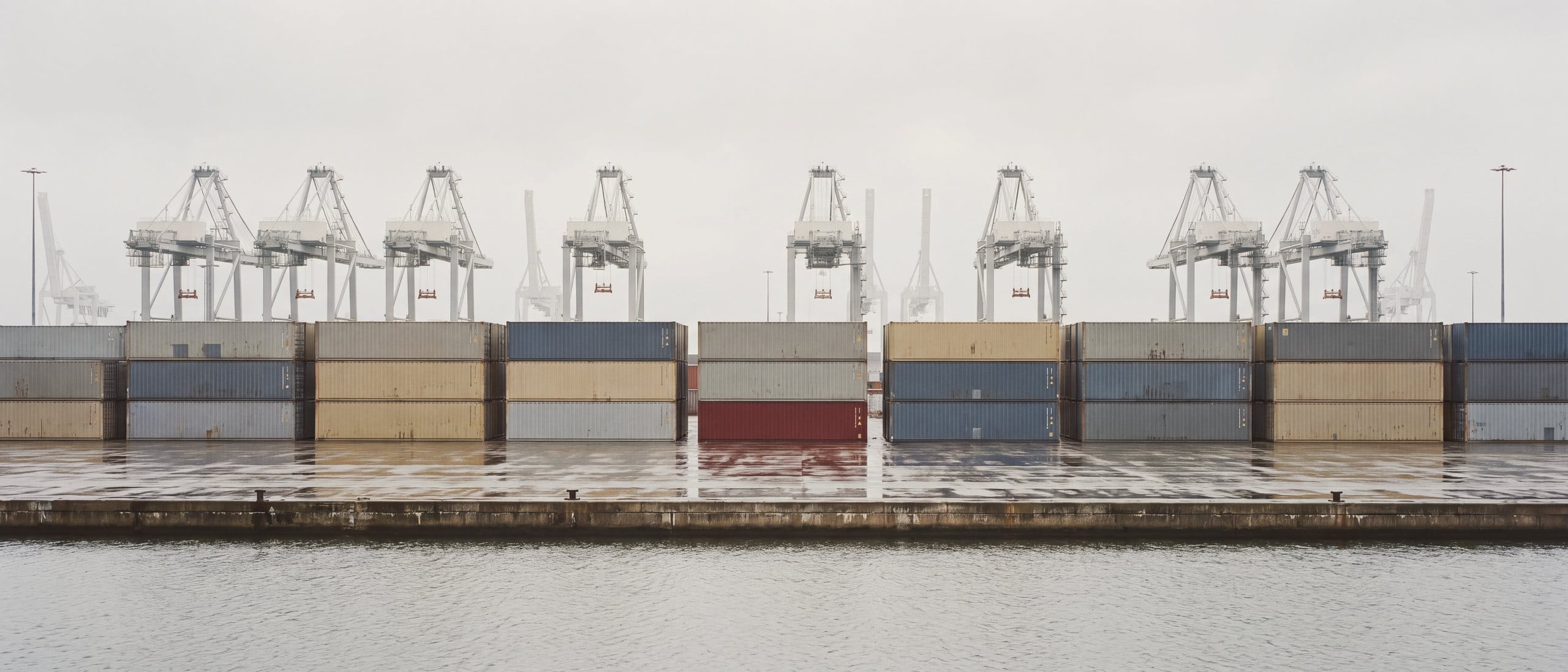 Container quays of a large North Sea port after rain, with quay cranes and one oxblood container among grey stacks.