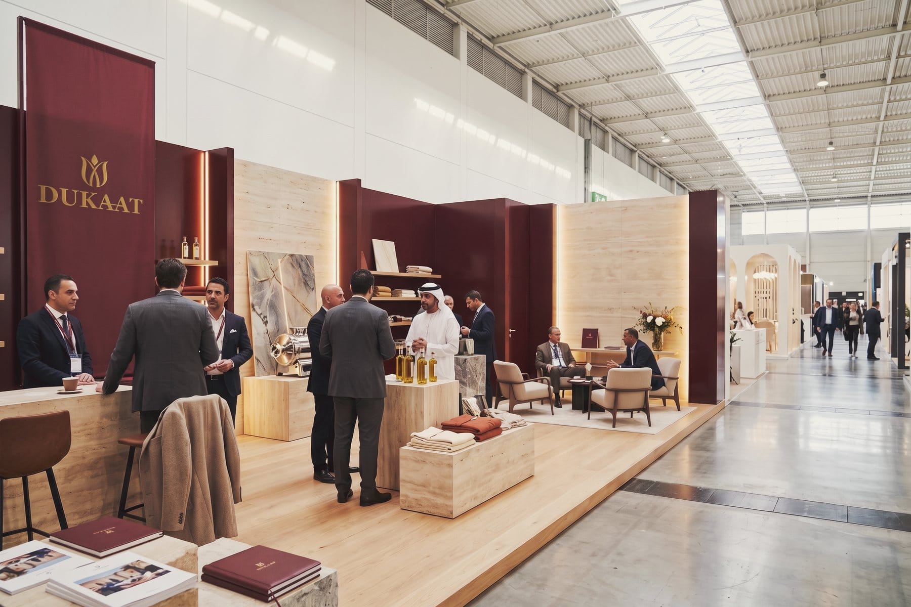 Het DUKAAT-paviljoen op een internationale beurs, met producten op stenen sokkels en bezoekers in gesprek.